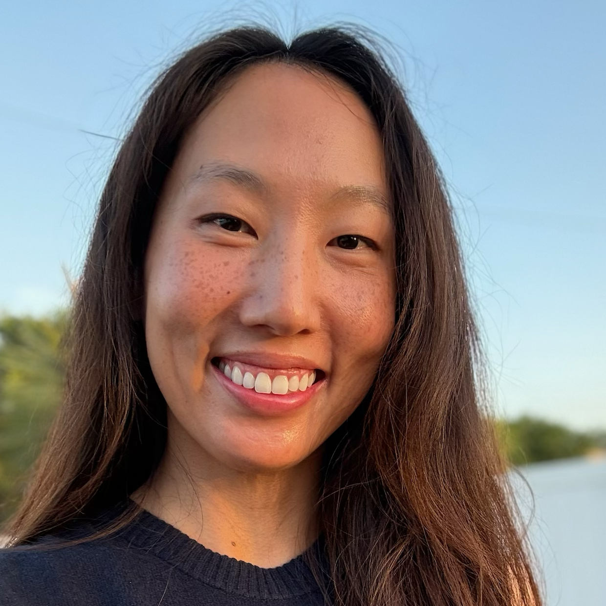 Headshot of a woman with long dark hair, smiling outdoors.