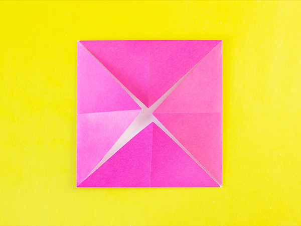 Pink origami square with folded corners on bright yellow background.