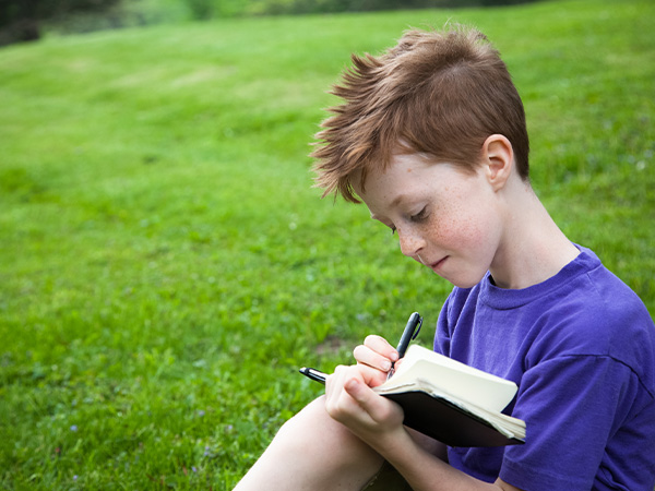 Young boy writing in notebook outdoors on grassy lawn.