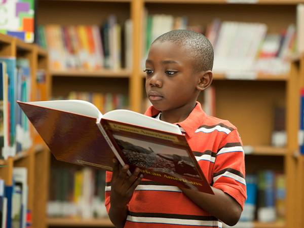 Young boy reading book in library with bookshelves in background.