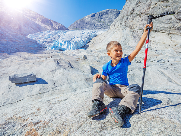 Young boy sitting on rocky terrain near glacier, holding hiking pole.