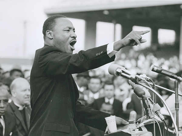 Martin Luther King, Jr., speaking and gesturing at a podium with microphones.