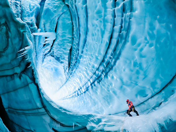 Ice cave explorer in red jacket amid swirling blue glacial formations.