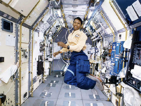 Astronaut floating in space station module wearing blue and beige uniform.