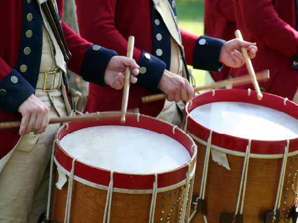 People in historical military uniforms playing large drums with drumsticks.