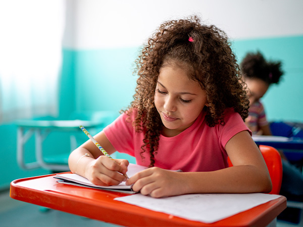 Curly-haired girl in pink shirt writing at school desk