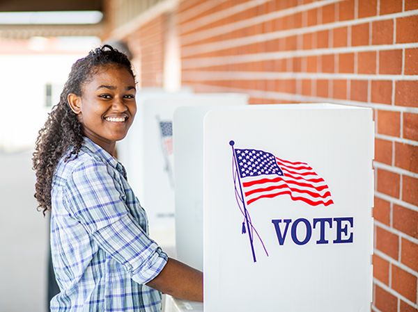 A smiling young woman stands next to a voting booth.