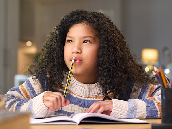 Here is simple alt text for this image in 10 words or less: Young girl with curly hair thinking while writing at desk