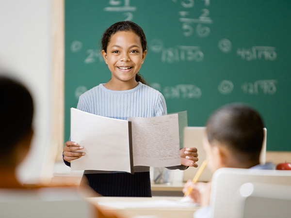 Smiling girl holding open book in front of chalkboard.