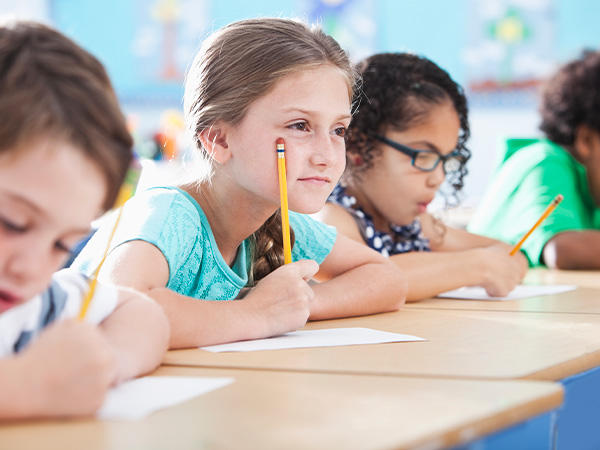 Three children sitting at a desk, one girl holding a pencil.