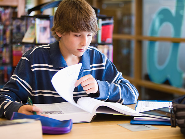 Young boy reading textbook in library or classroom setting.