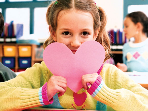 Smiling child holds pink paper heart in classroom.