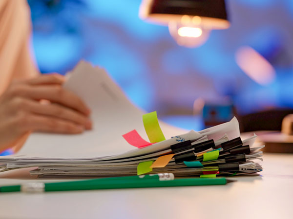 Person reviewing paperwork with colorful sticky tabs, green pencil on desk under lamp.